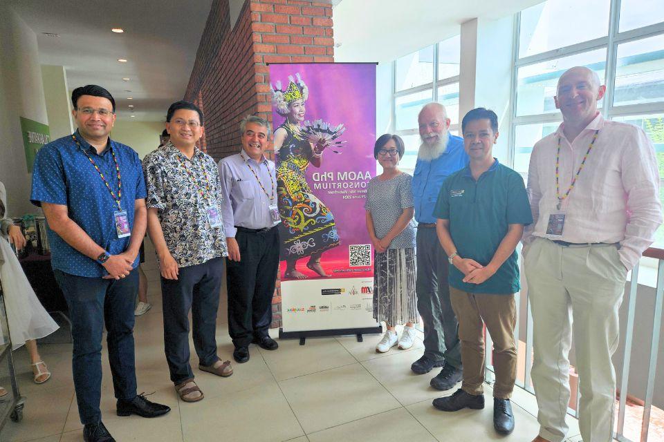 A group of conference attendees stand on either side of a banner that reads AAOM Ph.D. Consortium with an image of a Malaysian dancer in a colorful folk costume.